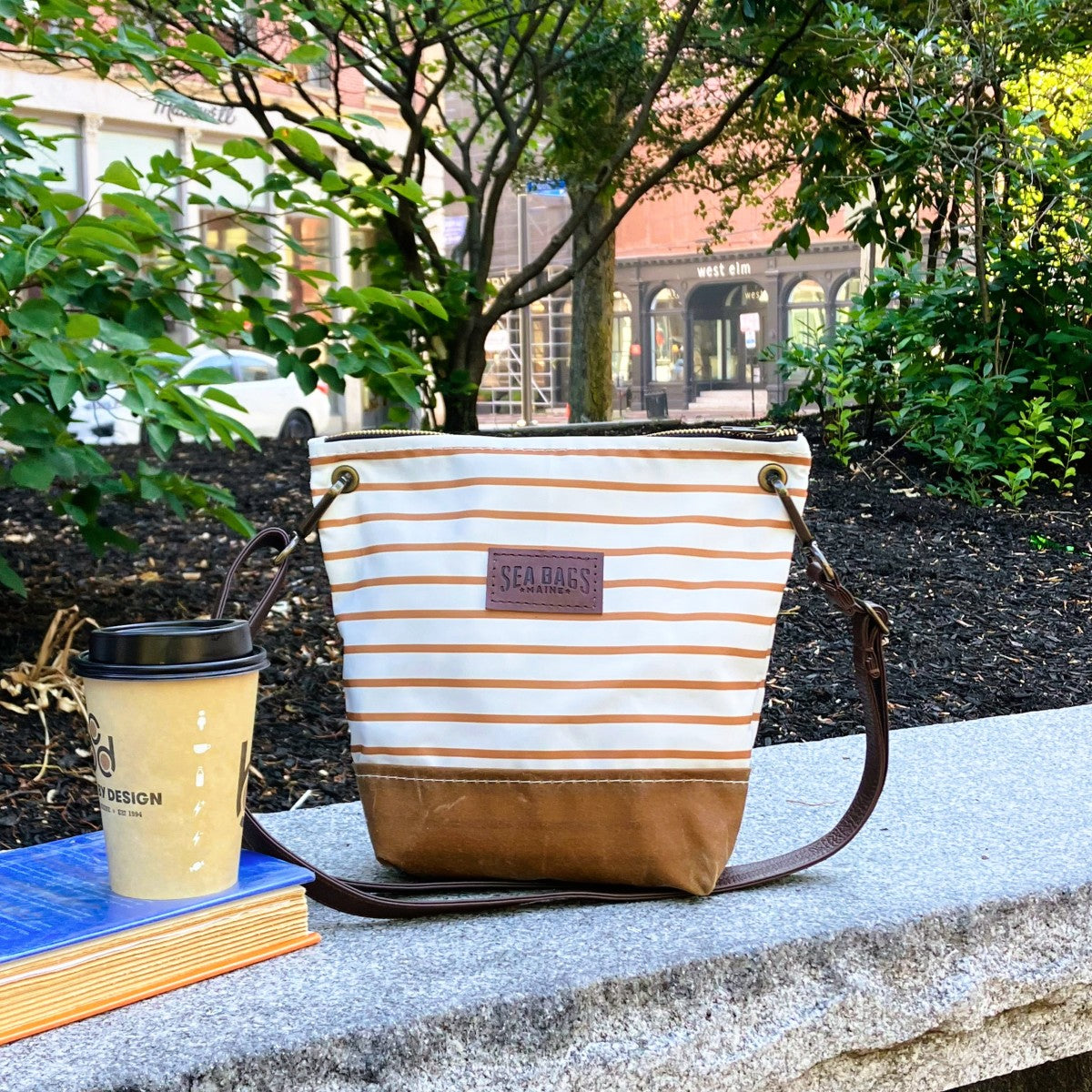 A Tan Chebeague Stripe Crossbody from Sea Bags, featuring a brown base and leather shoulder strap, sits on a stone bench beside a coffee cup and two books in an outdoor urban park setting. by Sea Bags