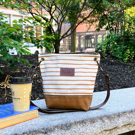 A Tan Chebeague Stripe Crossbody from Sea Bags, featuring a brown base and leather shoulder strap, sits on a stone bench beside a coffee cup and two books in an outdoor urban park setting. by Sea Bags
