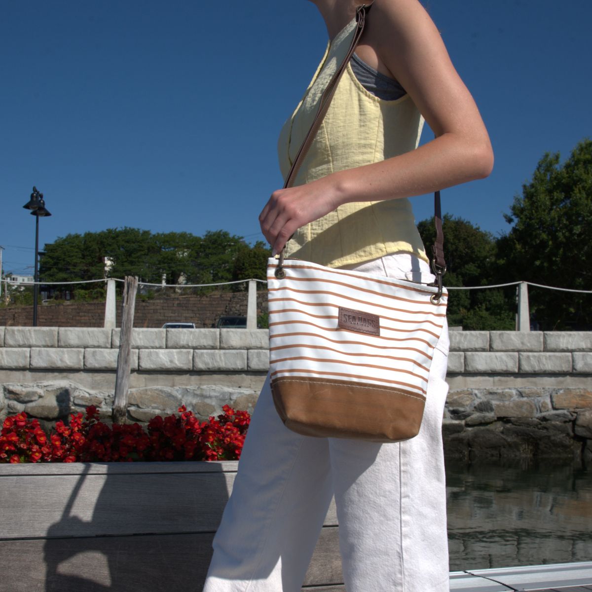A person wearing a yellow top and white pants carries the Sea Bags Tan Chebeague Stripe Crossbody, featuring a leather shoulder strap and crafted from recycled sailcloth, outdoors on a sunny day.