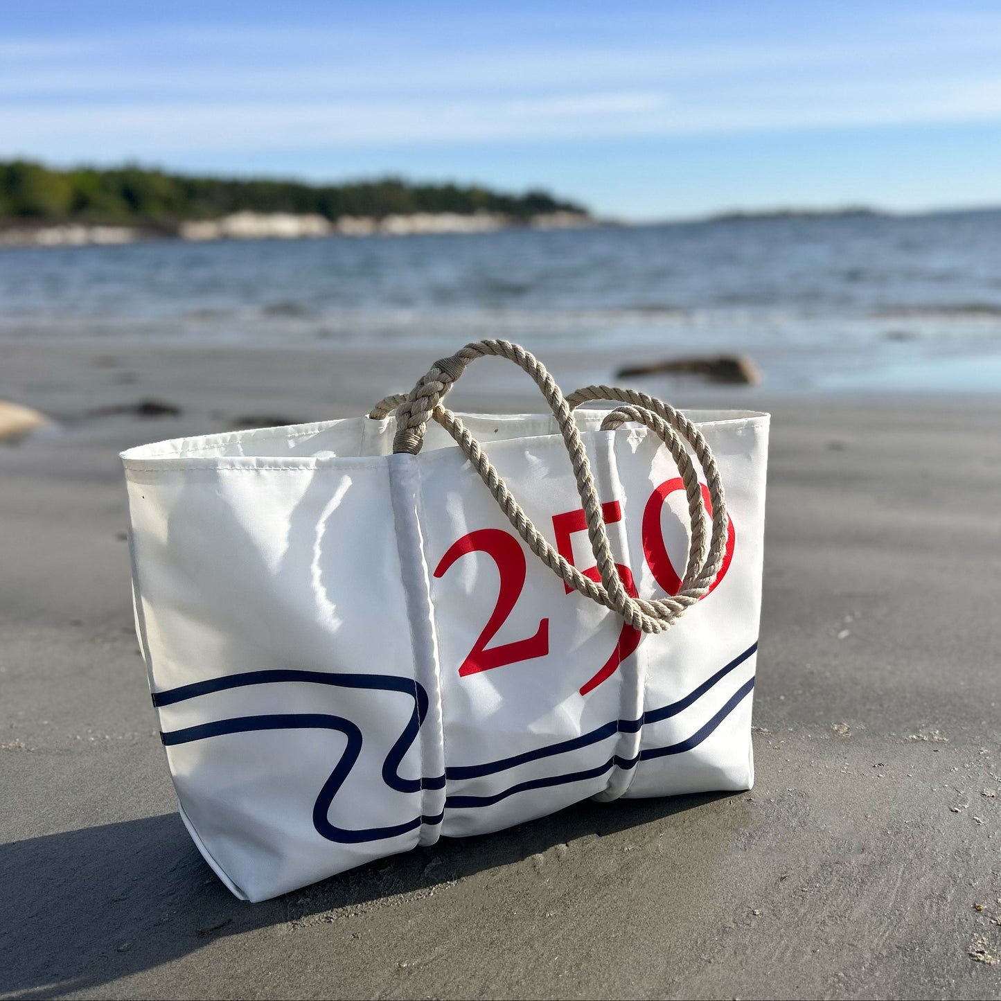 The Sea Bags 250th Patriotic Large Tote, crafted from recycled sail cloth and adorned with 250 in bold red, features sturdy rope handles and sits on a sandy beach with ocean and trees in the background. This handcrafted tote proudly showcases its Maine-made heritage. by Sea Bags
