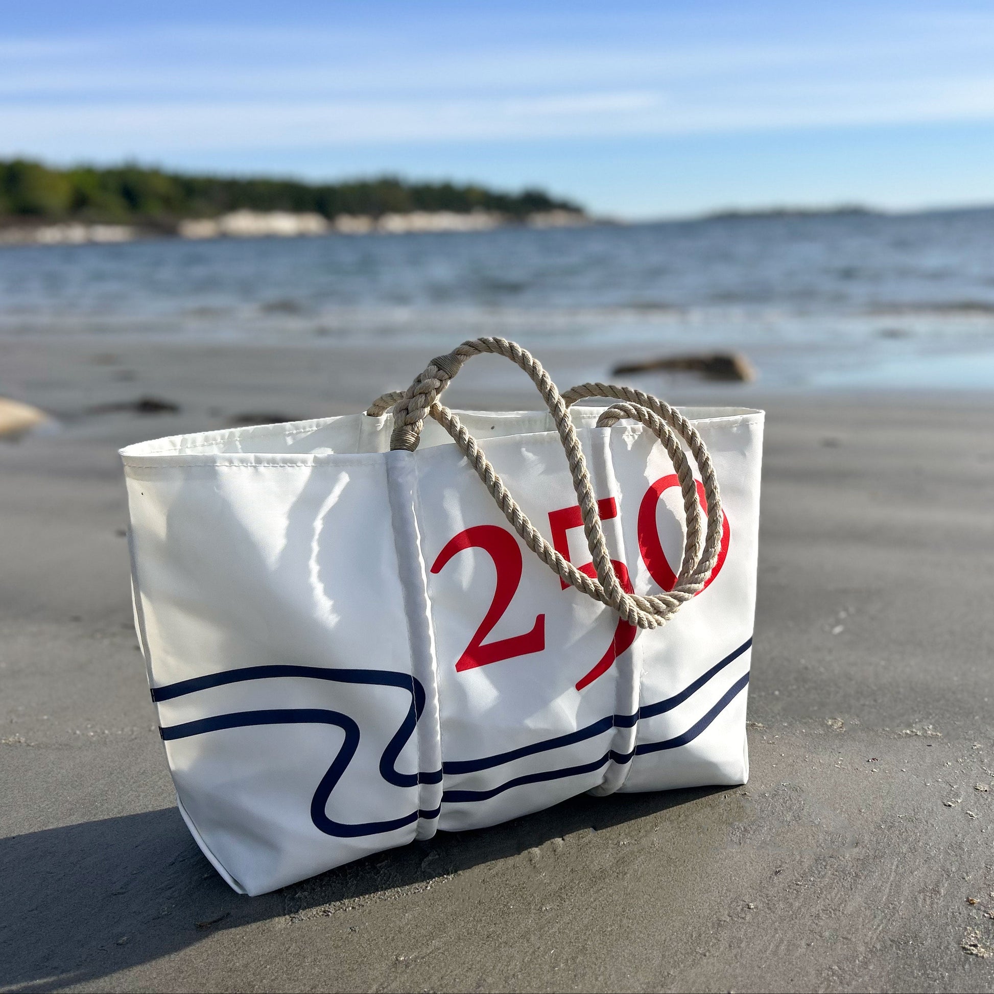 The Sea Bags 250th Patriotic Large Tote, crafted from recycled sail cloth and adorned with 250 in bold red, features sturdy rope handles and sits on a sandy beach with ocean and trees in the background. This handcrafted tote proudly showcases its Maine-made heritage. by Sea Bags