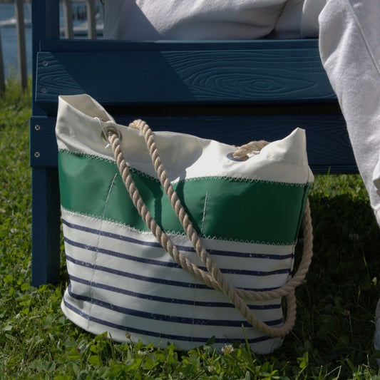 Sure Here’s the revised sentence using your product data: The Sea Bags Green-on-Navy Mariner Stripe Essential Handbag, crafted from lightweight recycled sail cloth, features green, white, and navy blue stripes with sturdy rope handles. Sitting on grass beside a person in white pants on a blue wooden bench, this eco-friendly tote combines everyday functionality with nautical-inspired style.
