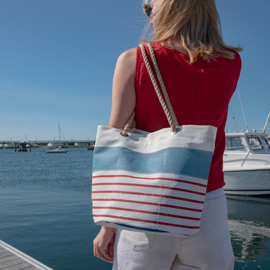 A woman viewed from behind stands at a waterfront wearing a large canvas tote bag with a white body, light blue horizontal stripe, and red horizontal stripes, plus thick rope handles. The background shows water, boats, and a blue sky.