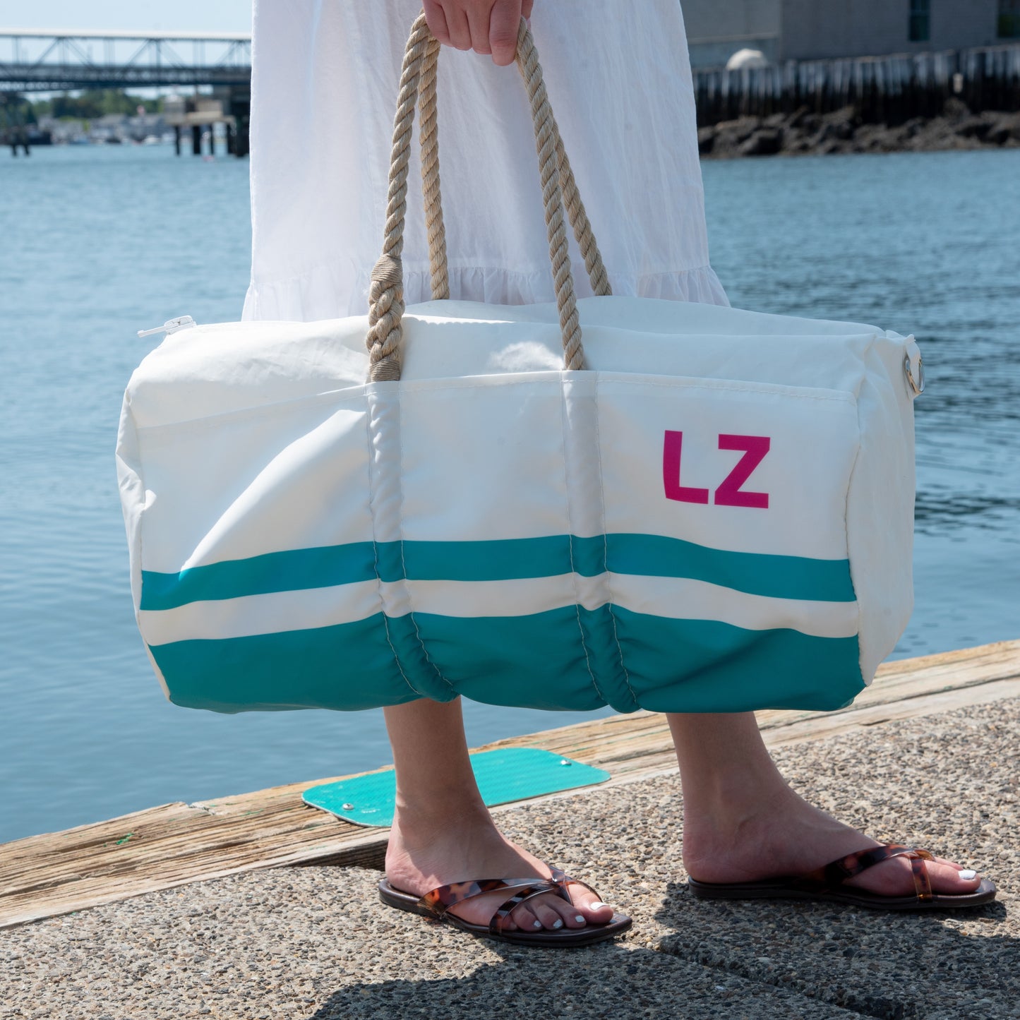 Wearing sandals and a white dress, a person stands on a dock by the water holding the Sea Bags Custom Monogram Breton Stripe Duffel  Bucket Bundle, crafted from recycled sailcloth and featuring the initials “LZ” printed on it.