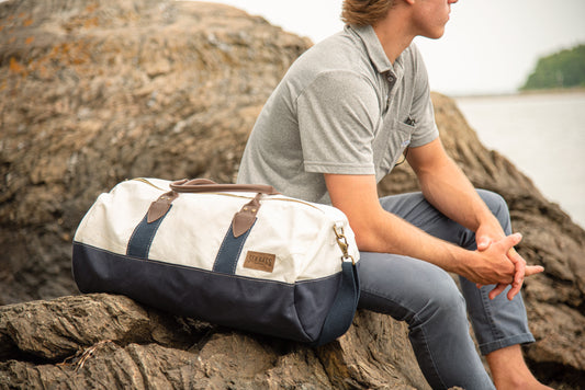 A person sits on rocks by the water, wearing a gray polo and blue pants, with the Sea Bags Chebeague Duffel—crafted from recycled sail and featuring premium leather handles—in white and navy beside them. by Sea Bags