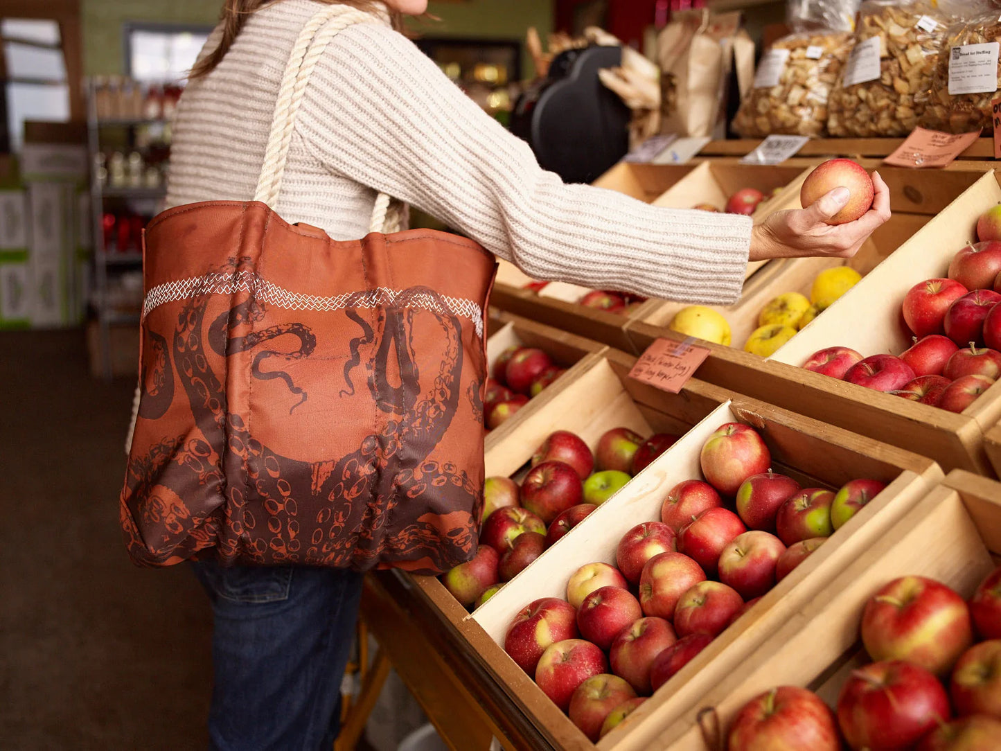 A shopper carries the Sea Bags Tanbark Octopus Medium Tote—crafted from recycled sail cloth and featuring an octopus design—while selecting apples in front of wooden bins at the market. by Sea Bags
