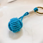 A Monkey Fist Key Ring - Turquoise by Mystic Knotwork, featuring a nautical monkey fist knot crafted from turquoise rope with white wrapping at the base and set on a brass ring, rests on a white surface.