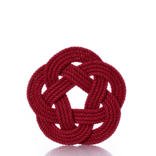 The true Nautical Sailor Knot Trivet - Red features a handwoven cotton rope, tied into an elaborate flat Turk’s head knot, reminiscent of classic nautical décor and set against a white background. by Sea Bags
