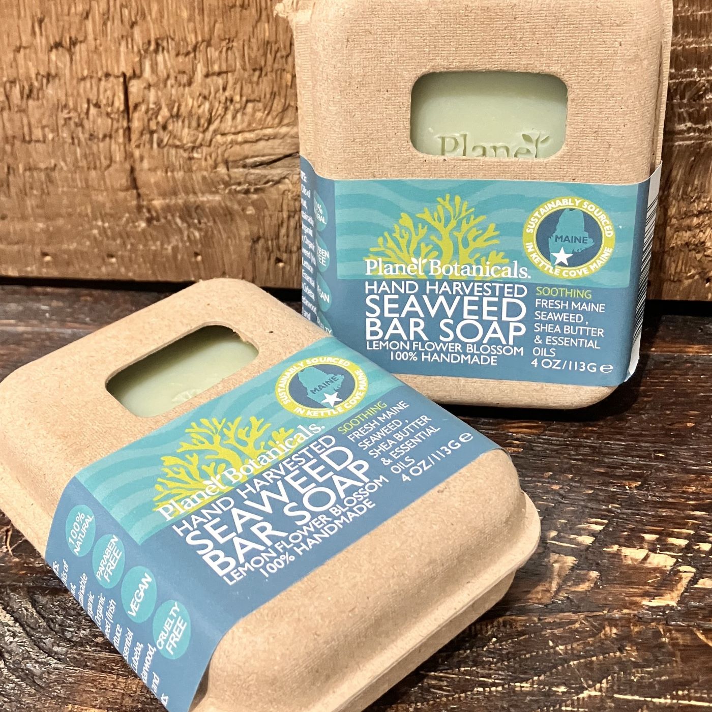 Two bars of Planet Botanicals Seaweed Bar Soap by True, hand-harvested and crafted with natural ingredients such as lemon flower blossom, are displayed in eco-friendly packaging on a wooden surface.
