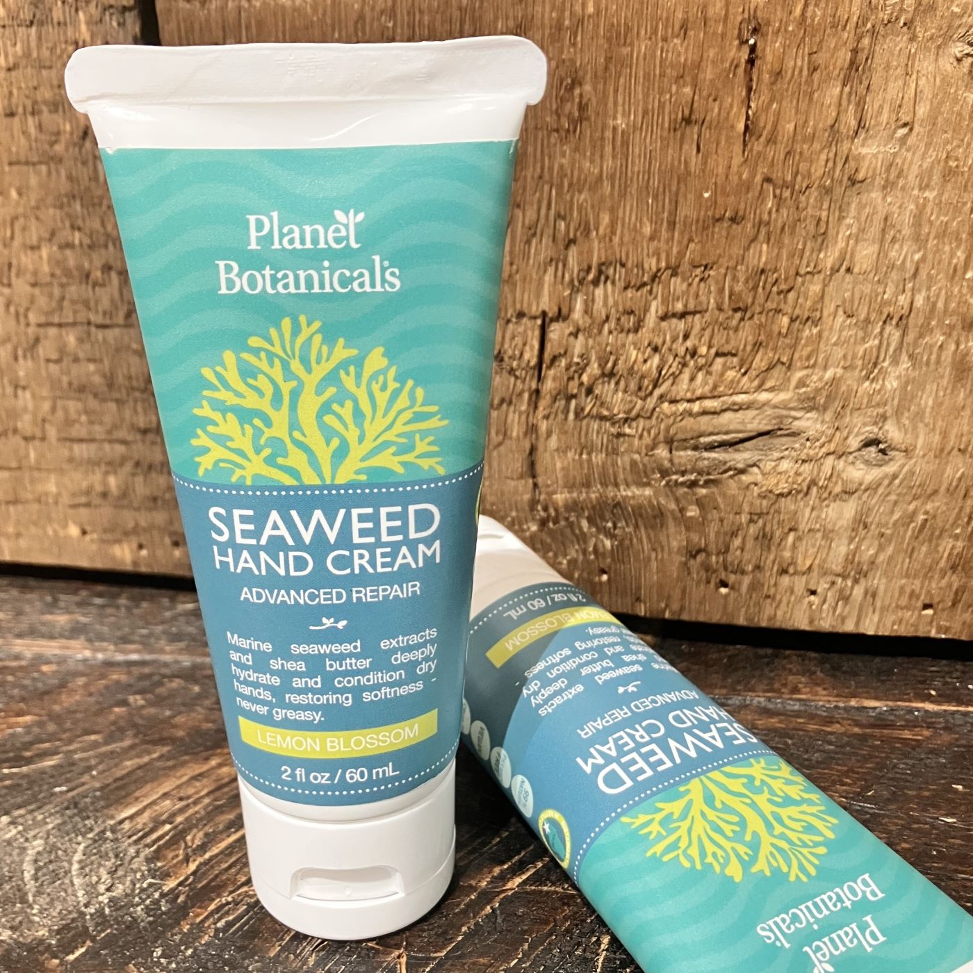 A tube of Planet Botanicals Seaweed Hand Cream 2 oz stands upright, with another tube lying beside it on a wooden surface—both showcasing the luxurious skincare benefits of this Maine-made hand cream from Planet Botanicals.