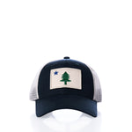 The Maine Flag Trucker Hat by true features a navy blue and white mesh design with a front patch depicting a green tree and blue star on a beige background, offering classic New England style inspired by the iconic Maine state flag.