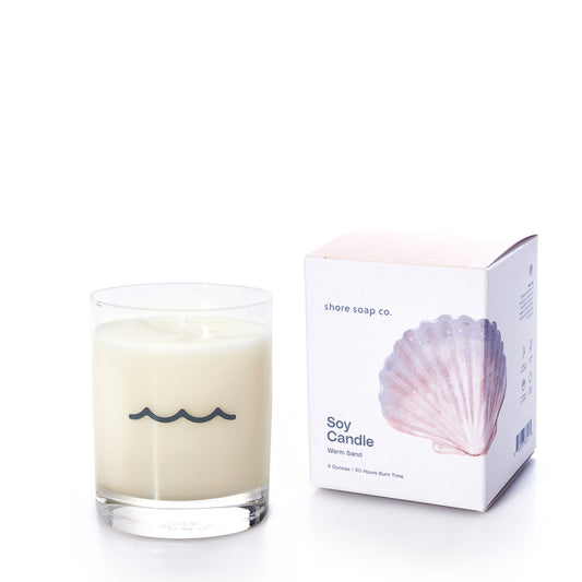 The Warm Sand Eco-Friendly Candle by Shore Soap Co features a white soy wax candle in a reusable tumbler glass adorned with a wavy line, accompanied by a box decorated with a seashell image and Shore Soap Co branding. This candle fills your space with the soothing Warm Sand fragrance.