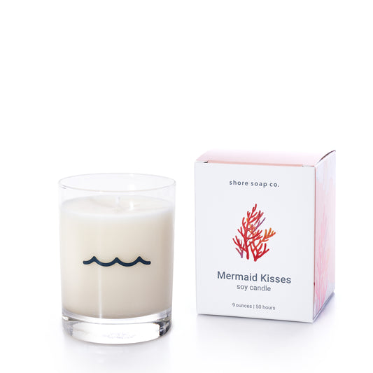 A white Mermaid Kisses Eco-Friendly Candle from Shore Soap Co, crafted from clean-burning soy wax, sits in a clear glass with a wave design next to a box labeled Mermaid Kisses and adorned with a red coral illustration—an elegant addition to Shore Soap Co’s collection of handcrafted personal care products.