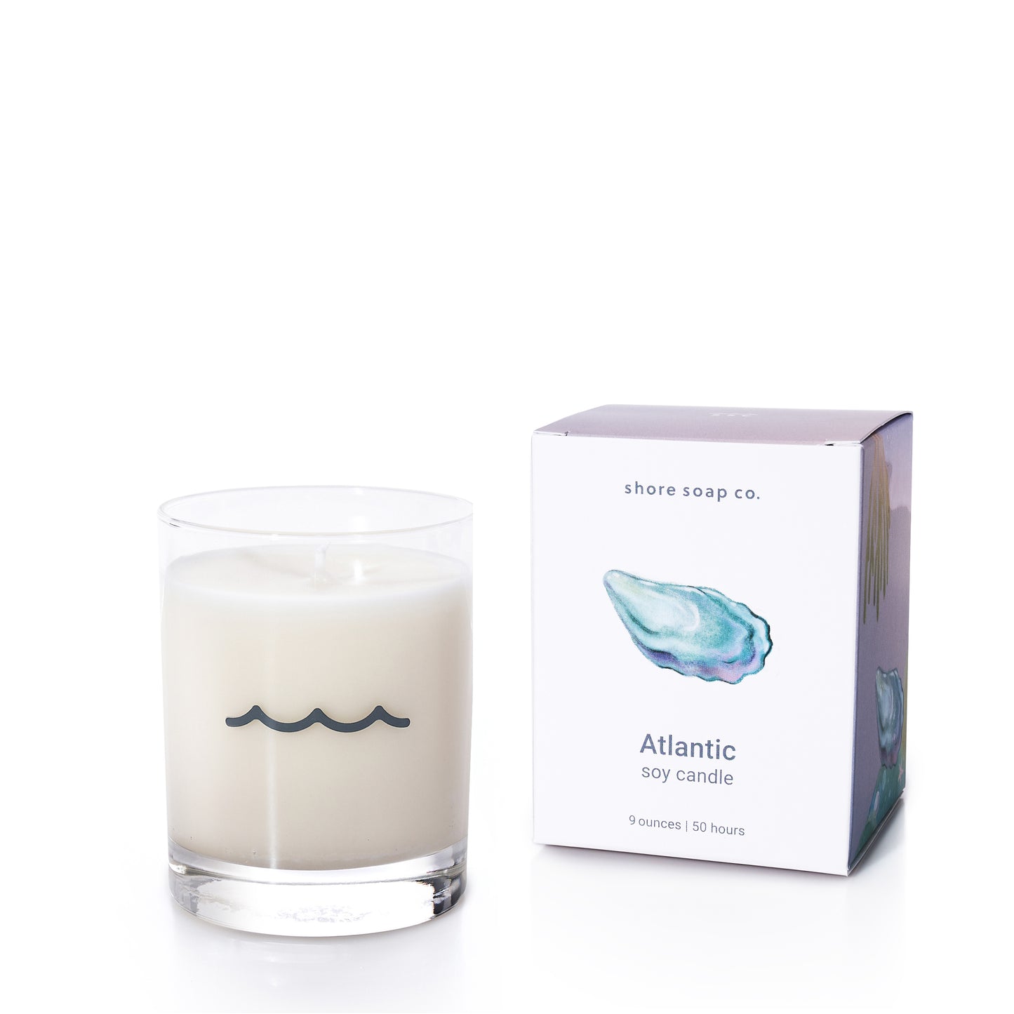 The Atlantic Eco-Friendly Candle by Shore Soap Co.—a white soy wax candle in a clear, reusable tumbler glass adorned with a wave symbol—rests beside its box featuring an oyster shell illustration and fills the air with the inviting Warm Sand scent.