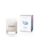 The Atlantic Eco-Friendly Candle by Shore Soap Co.—a white soy wax candle in a clear, reusable tumbler glass adorned with a wave symbol—rests beside its box featuring an oyster shell illustration and fills the air with the inviting Warm Sand scent.