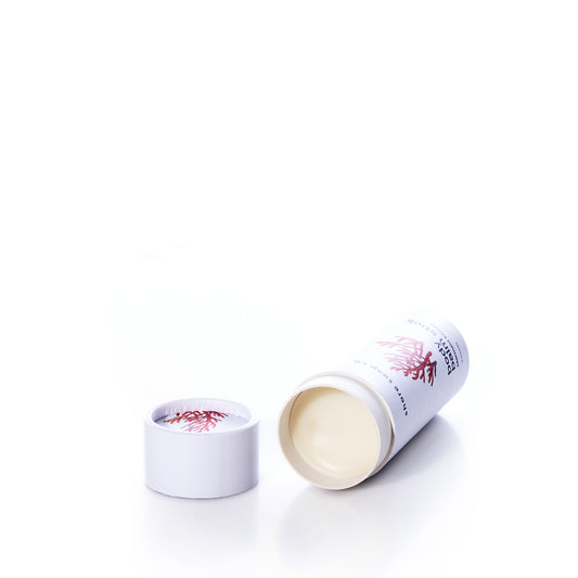A white cylindrical container of Shore Soap Co’s Mermaid Kisses Eco-Friendly Body Balm rests on its side with the lid removed, revealing the balm inside. The container features minimal red and black text and graphics, highlighting this natural moisturizer from Shore Soap Co’s line of handcrafted personal care products. by Sea Bags