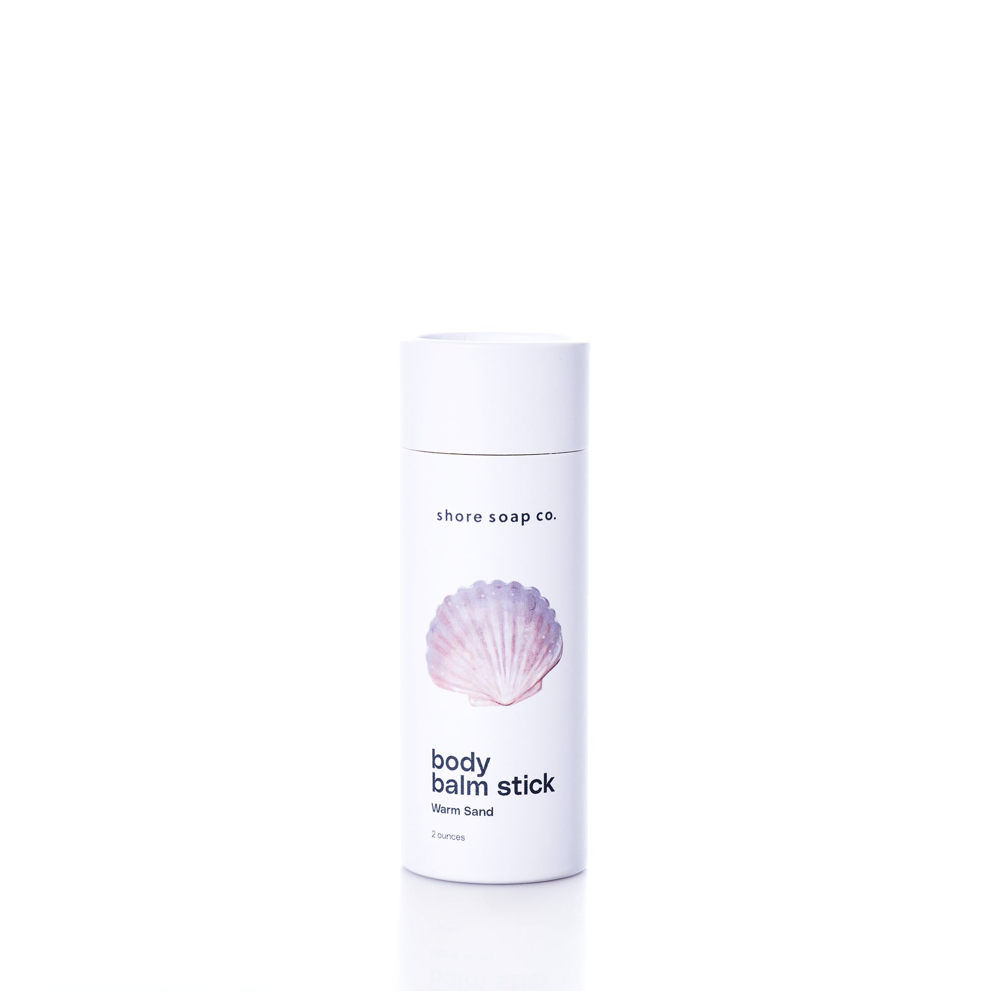 A white cylindrical container labeled Shore Soap Co Warm Sand Eco-Friendly Body Balm Stick, featuring a pink seashell graphic on the front, stands on a white background; this concentrated body balm delivers lasting hydration for your skin.