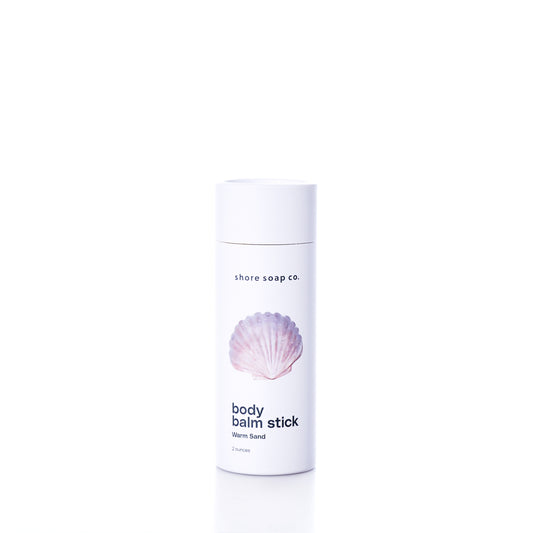 A white cylindrical container labeled Shore Soap Co Warm Sand Eco-Friendly Body Balm Stick, featuring a pink seashell graphic on the front, stands on a white background; this concentrated body balm delivers lasting hydration for your skin.