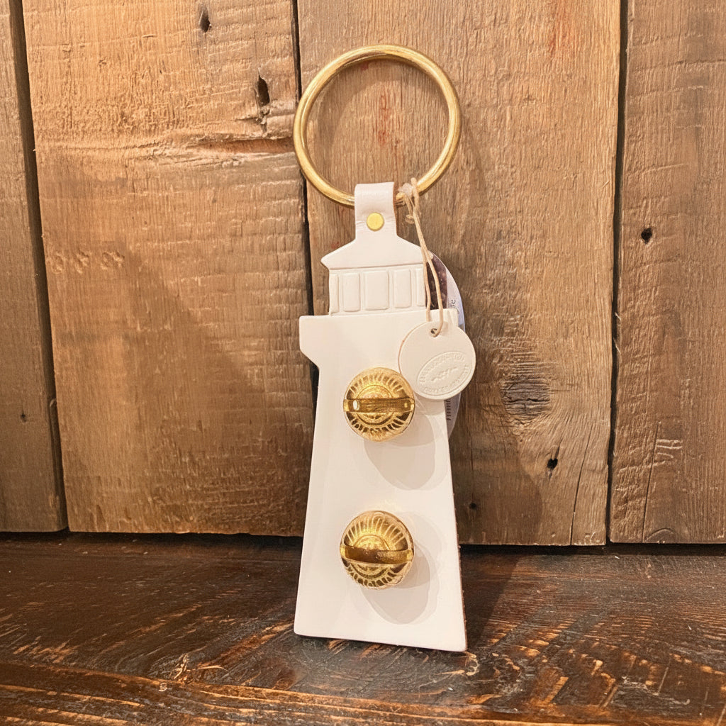 White Lighthouse Door Chime by New England Bells, featuring a classic lighthouse silhouette in white with a gold keyring and two decorative gold knobs, displayed upright on a wooden surface against a wood panel backdrop—evoking the timeless charm characteristic of New England Bells’ handcrafted designs.
