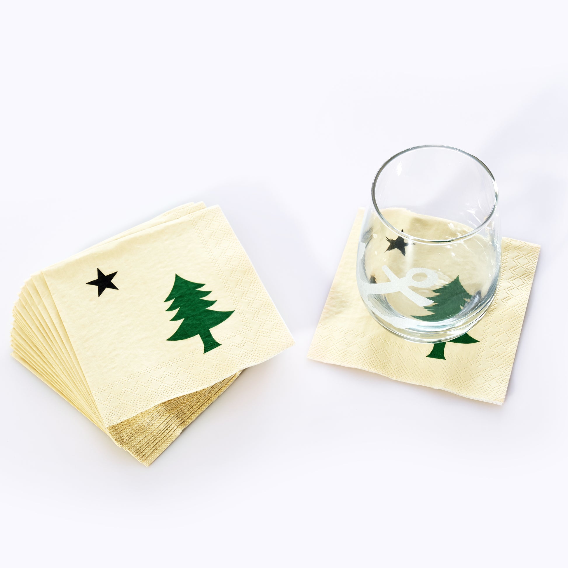 A glass rests on a Maine Bicentennial Cocktail Napkin by Boston International, decorated with a green Christmas tree and black star reminiscent of the pine-tree-and-star flag, while a stack of identical napkins sits nearby. by Sea Bags