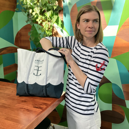 A woman wearing the Batela USA Inc 3/4 Sleeve Boatneck Top in Navy Breton Stripe  Red Anchor stands indoors, reaching into a white and navy nautical tote bag placed on a wooden table. A green leafy plant and a colorful wall are visible in the background.