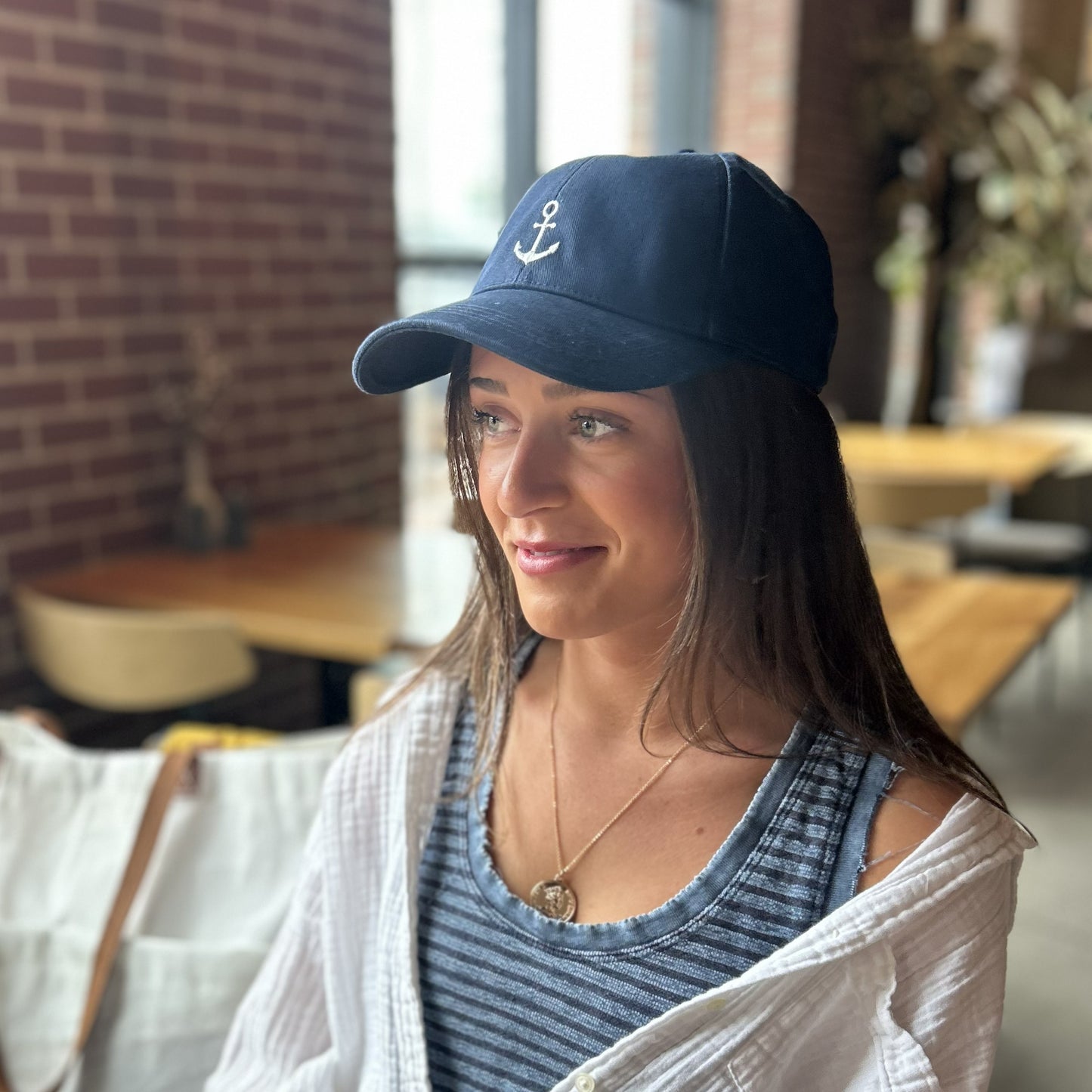 A woman wearing the Anchor Baseball Hat - Navy by true, along with a striped tank top and a white shirt, sits indoors at a wooden table with a blurred background.