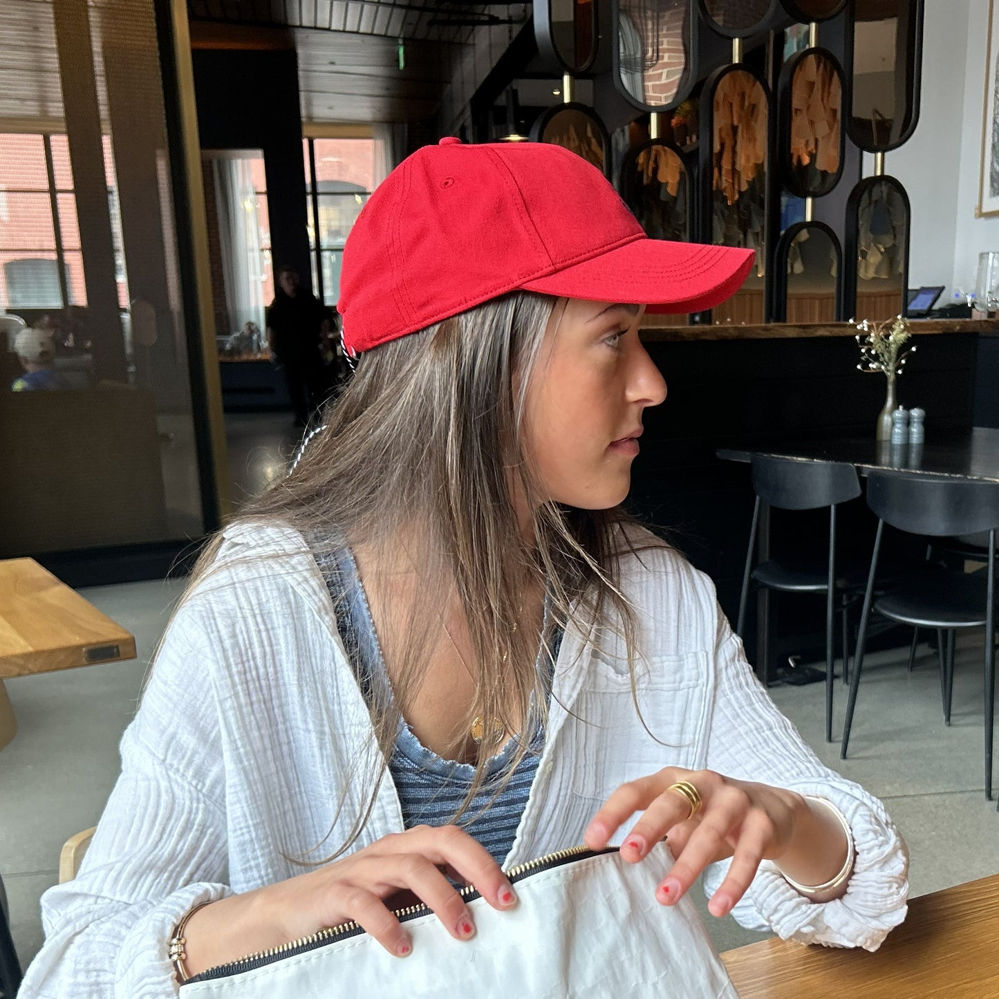 A woman wearing the Batela USA Inc Anchor Baseball Hat in red and a white shirt sits at a table in a modern café, holding a white bag and looking to her left—her style perfectly suited for nautical outings. by Sea Bags