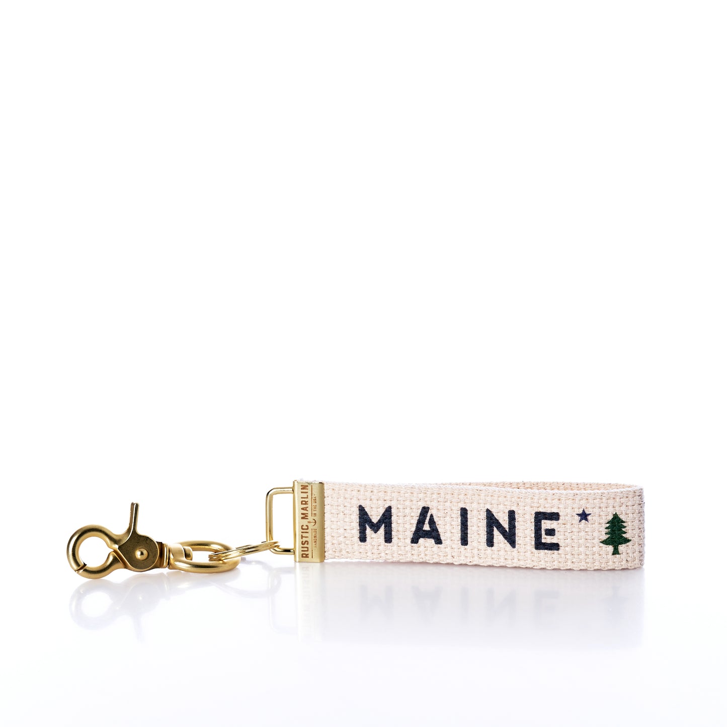 Introducing the Rustic Marlin - Keychain Maine Bicentennial by Rustic Marlin: a beige fabric keychain with a gold clasp, embroidered with MAINE and a small green pine tree graphic—perfect as a Maine Pine Tree bag charm or stylish key ring. by Sea Bags