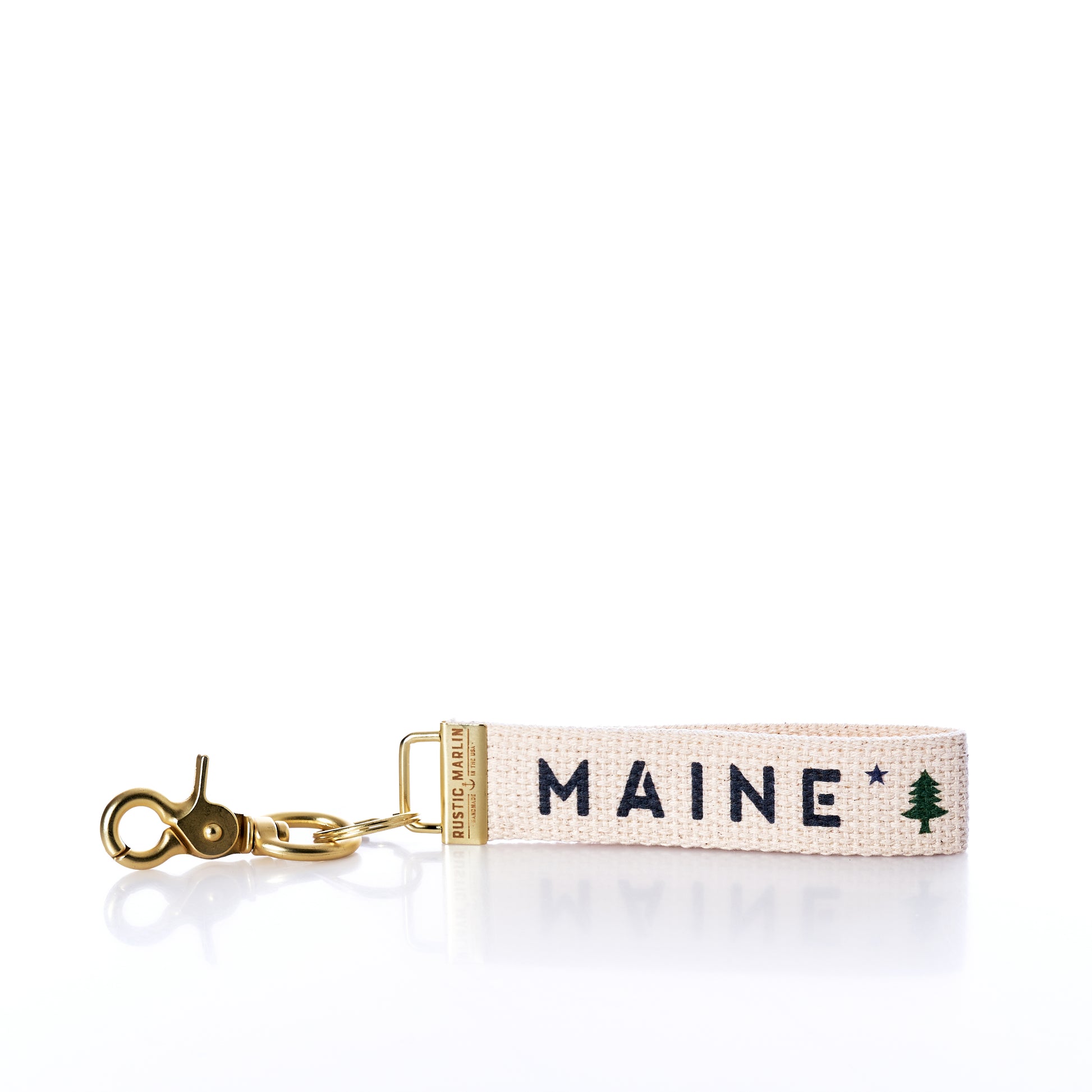 Introducing the Rustic Marlin - Keychain Maine Bicentennial by Rustic Marlin: a beige fabric keychain with a gold clasp, embroidered with MAINE and a small green pine tree graphic—perfect as a Maine Pine Tree bag charm or stylish key ring. by Sea Bags
