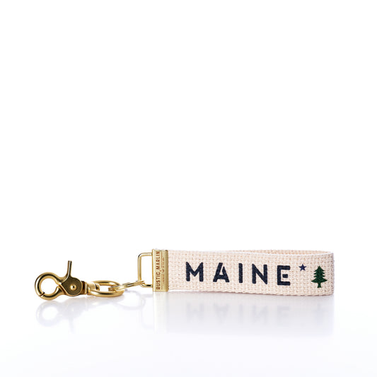 Introducing the Rustic Marlin - Keychain Maine Bicentennial by Rustic Marlin: a beige fabric keychain with a gold clasp, embroidered with MAINE and a small green pine tree graphic—perfect as a Maine Pine Tree bag charm or stylish key ring. by Sea Bags