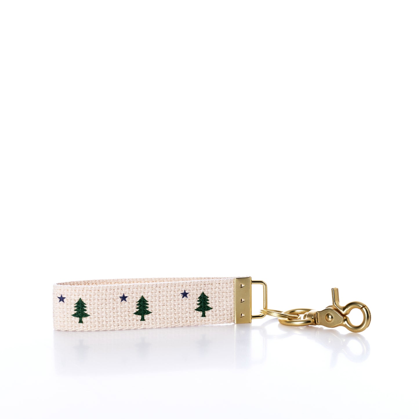 The Rustic Marlin - Keychain Maine Bicentennial by Rustic Marlin is a beige fabric keychain adorned with Maine Pine Tree and blue star motifs. It features gold metal hardware and a swivel clasp, making it perfect as a distinctive bag charm or key ring. Shown against a white background. by Sea Bags