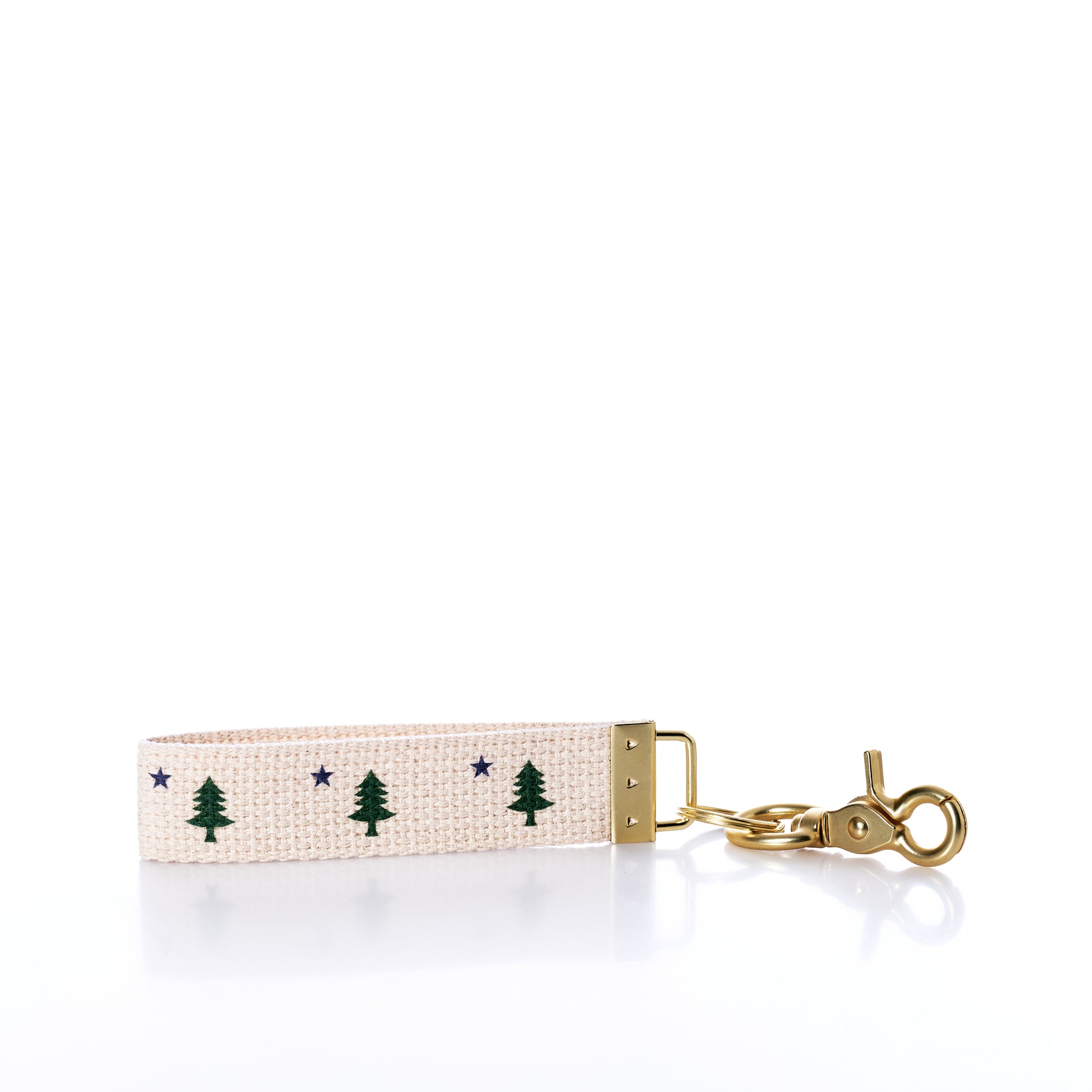 The Rustic Marlin - Keychain Maine Bicentennial by Rustic Marlin is a beige fabric keychain adorned with Maine Pine Tree and blue star motifs. It features gold metal hardware and a swivel clasp, making it perfect as a distinctive bag charm or key ring. Shown against a white background. by Sea Bags