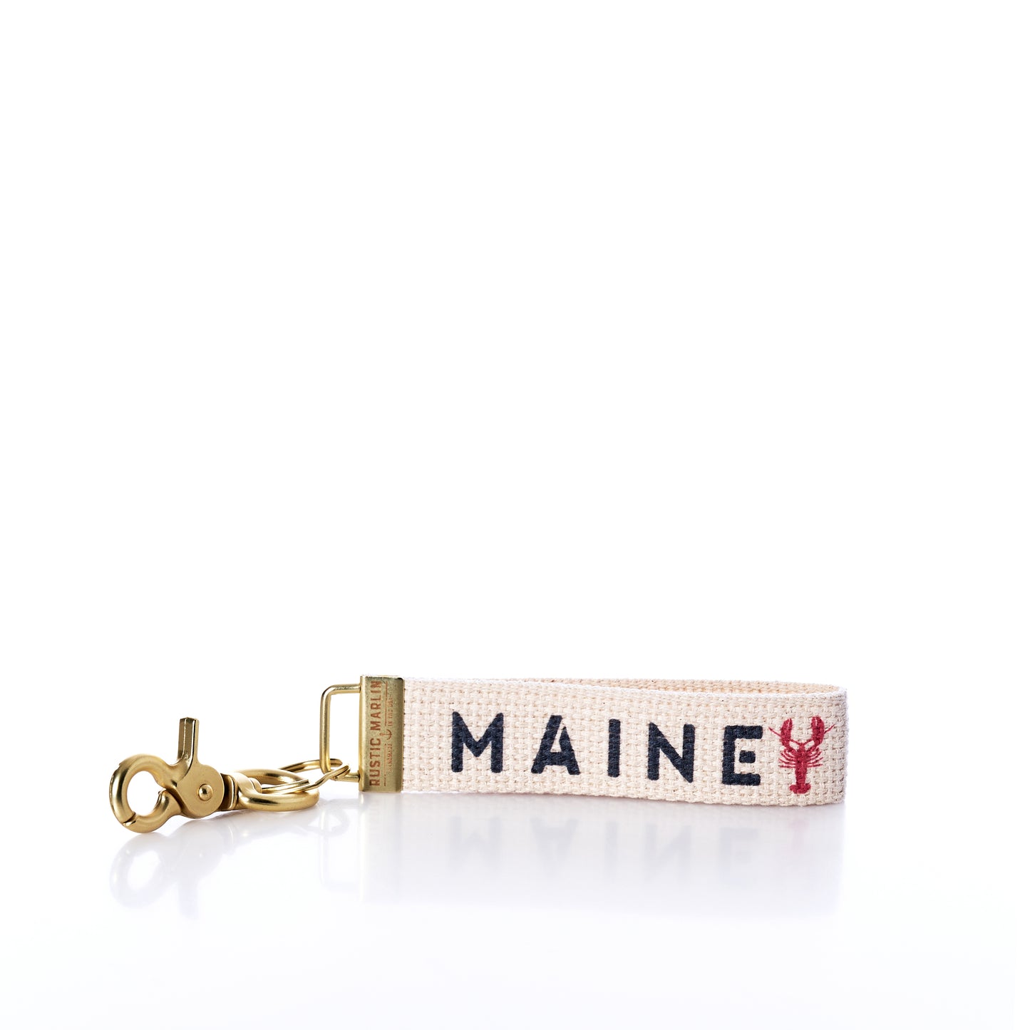 The Rustic Marlin - Keychain Maine Lobsters by Rustic Marlin is a woven bag charm keychain featuring gold hardware and reads MAINE in blue letters, with a small red lobster symbol replacing the letter I on a white background—making it a delightful Maine souvenir for any lobster enthusiast. by Sea Bags