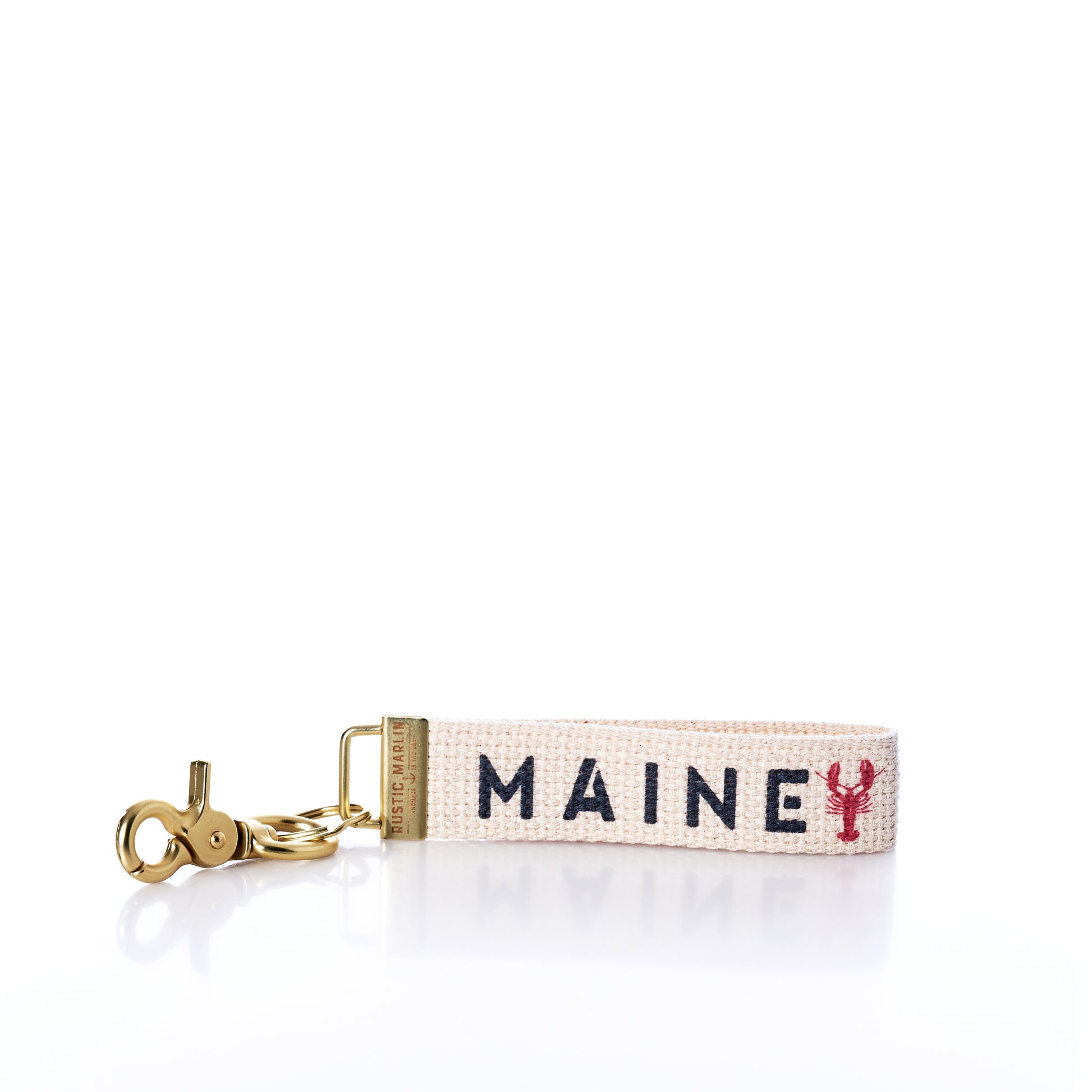The Rustic Marlin - Keychain Maine Lobsters by Rustic Marlin is a woven bag charm keychain featuring gold hardware and reads MAINE in blue letters, with a small red lobster symbol replacing the letter I on a white background—making it a delightful Maine souvenir for any lobster enthusiast. by Sea Bags