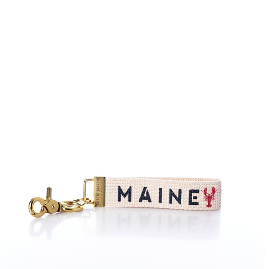 The Rustic Marlin - Keychain Maine Lobsters by Rustic Marlin is a woven bag charm keychain featuring gold hardware and reads MAINE in blue letters, with a small red lobster symbol replacing the letter I on a white background—making it a delightful Maine souvenir for any lobster enthusiast. by Sea Bags
