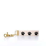 The Rustic Marlin - Keychain Paw Prints by Rustic Marlin features a beige fabric design with three black paw prints and a gold metal clasp, making it a stylish bag charm, displayed on a white background. by Sea Bags