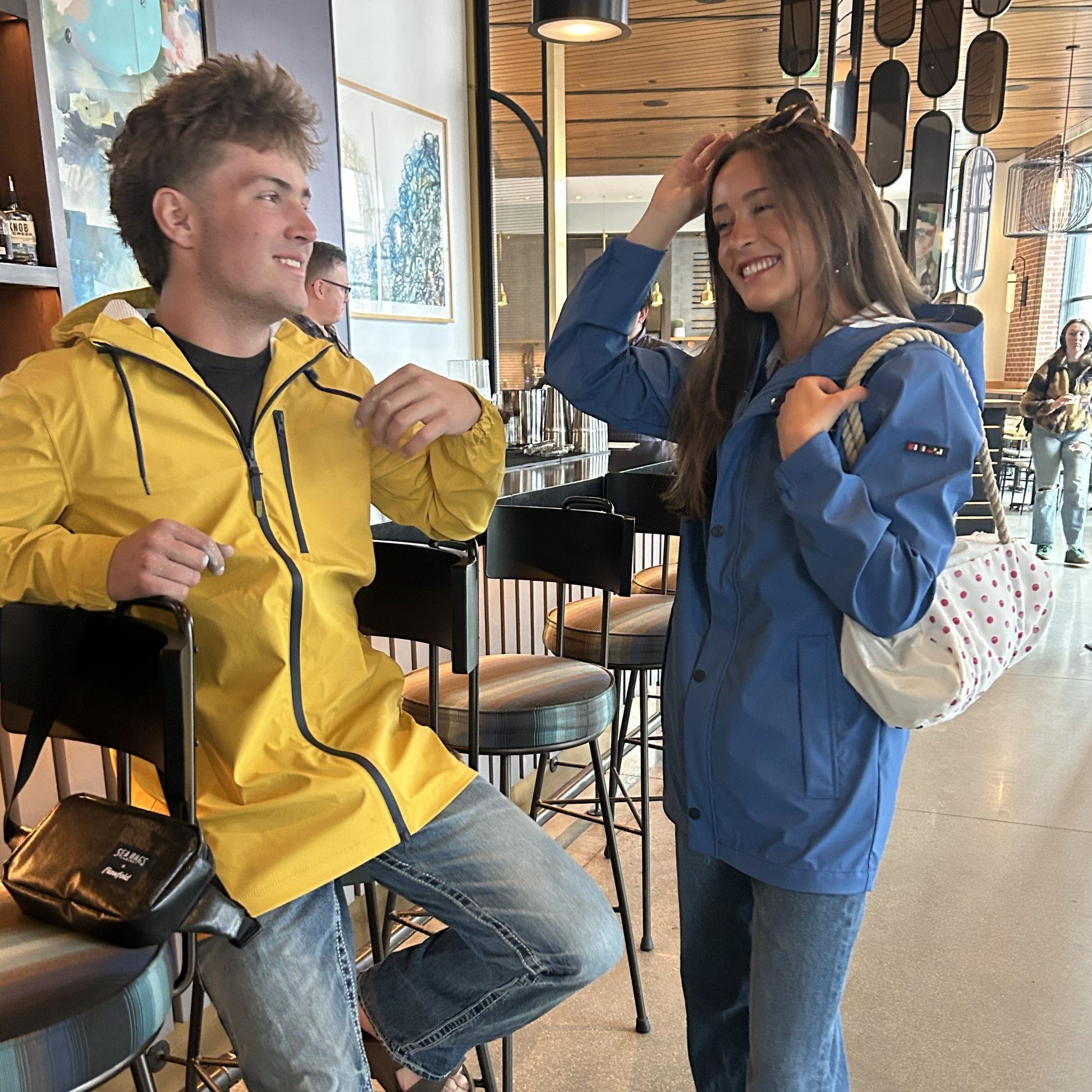 Two people in waterproof shells, one wearing the Batela USA Inc Harborside Waterproof Raincoat in yellow and the other in blue, are talking and smiling inside a modern café. Other patrons and bar stools are visible in the background. by Sea Bags