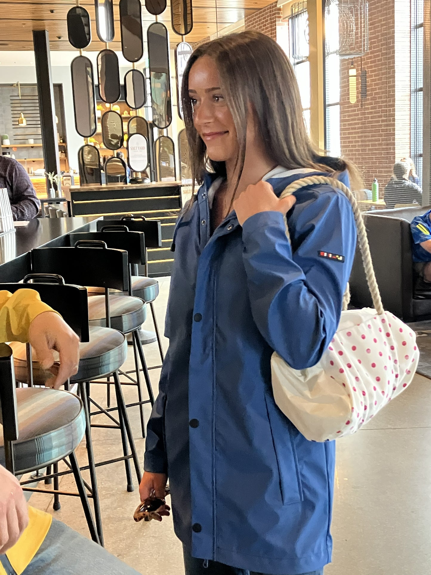 A woman wearing the Harborside Waterproof Raincoat in High Tide by Batela USA Inc stands indoors, holding a white bag with red polka dots over her shoulder. She is facing another person and appears to be in a modern cafe or restaurant setting, perhaps taking a break from coastal adventures. by Sea Bags