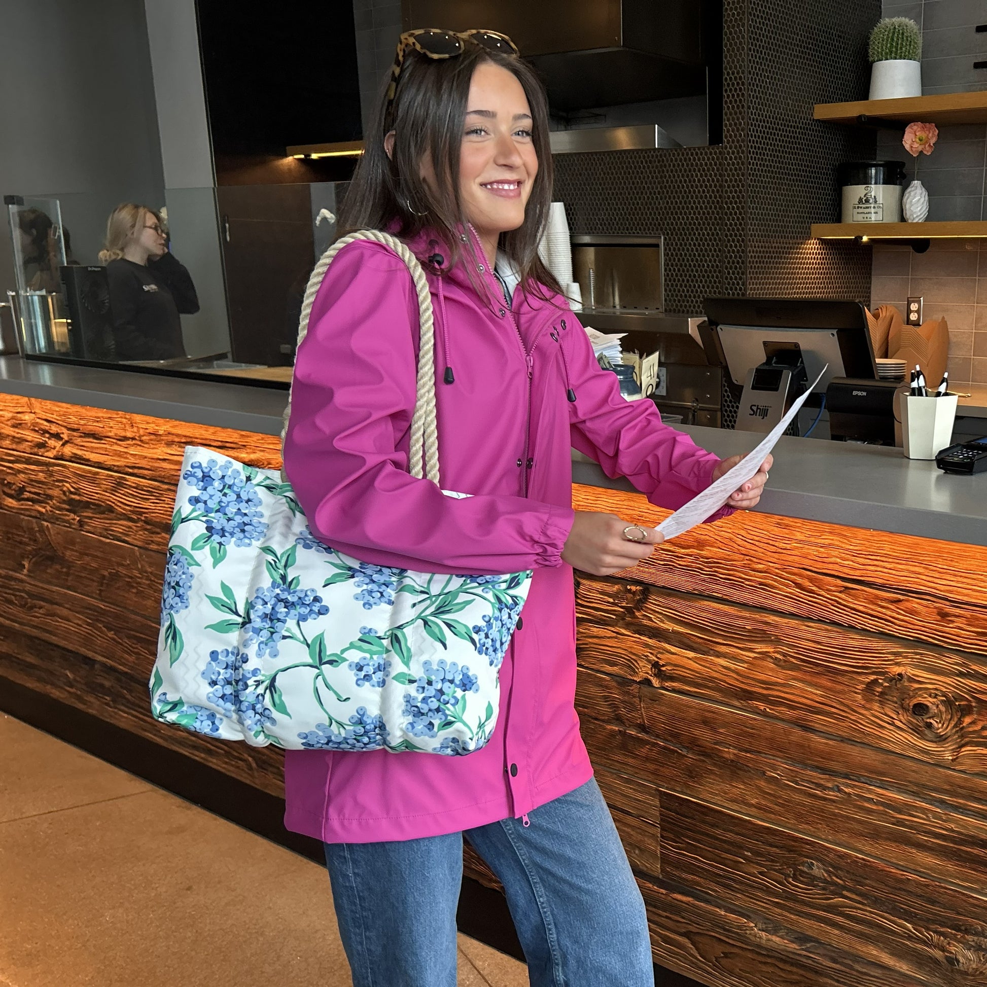 A woman in a bright pink jacket stands at a reception desk, holding a paper and the Sea Bags Blueberry Print Handbag—crafted from recycled sail cloth and adorned with Maine blueberries—while smiling. by Sea Bags