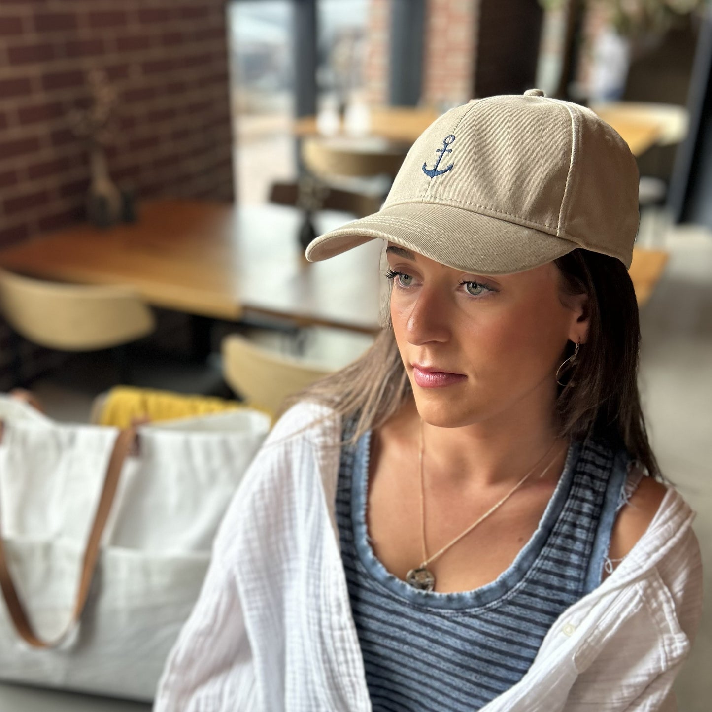 Wearing the Batela USA Inc Anchor Baseball Hat in beige and a striped shirt, a woman sits indoors near a wooden table, gazing thoughtfully ahead. A large white bag in the background hints at her appreciation for both style and sun protection. by Sea Bags