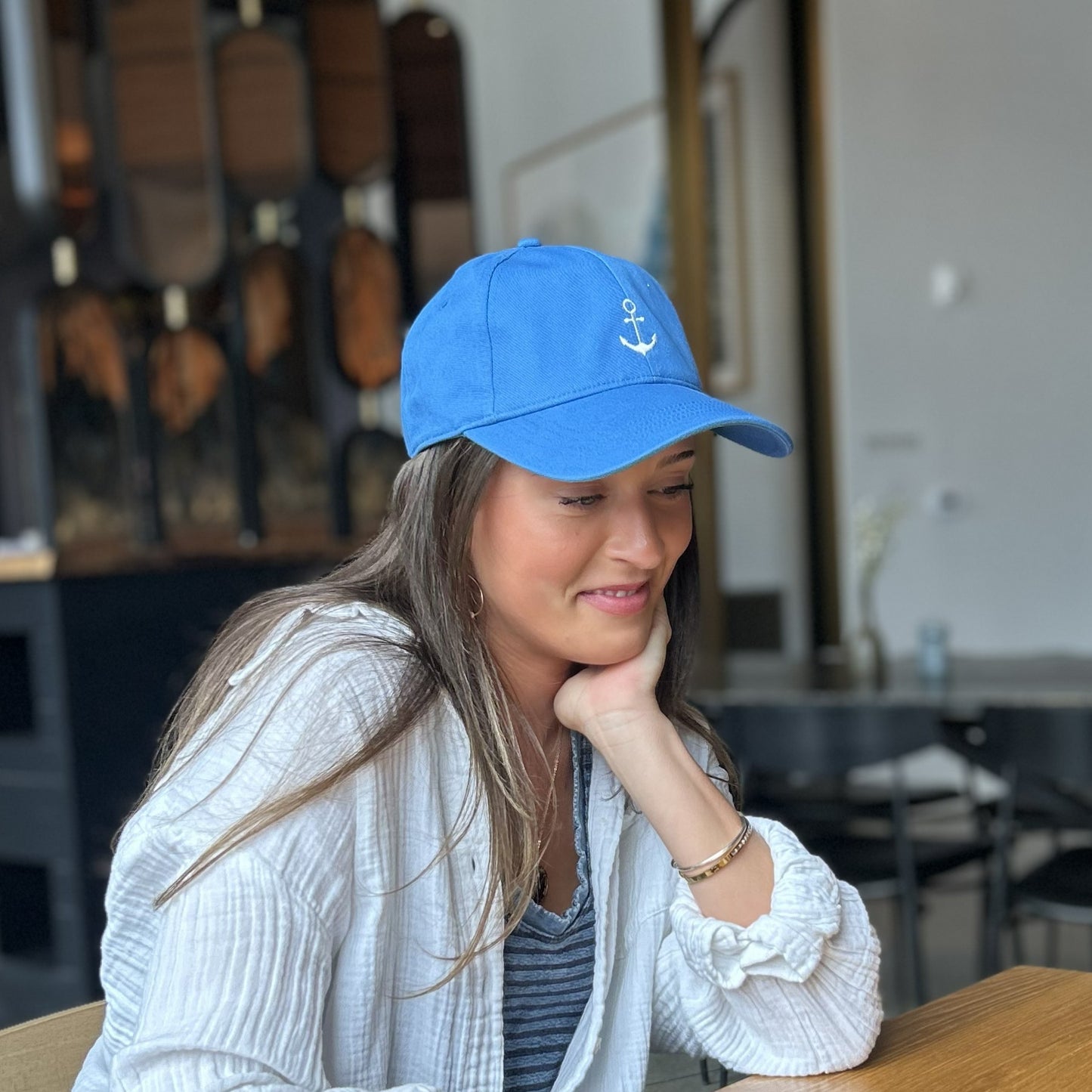 A woman sits indoors at a table, smiling and looking down, wearing the Batela USA Inc Anchor Baseball Hat - High Tide with an embroidered anchor symbol, paired with a white shirt and striped top. by Sea Bags