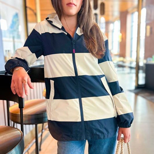 A woman wearing the Harborside Waterproof Raincoat in Stripe Navy  White by true stands indoors, leaning on a chair and holding a rope strap bag, with blurred tables and windows in the background.
