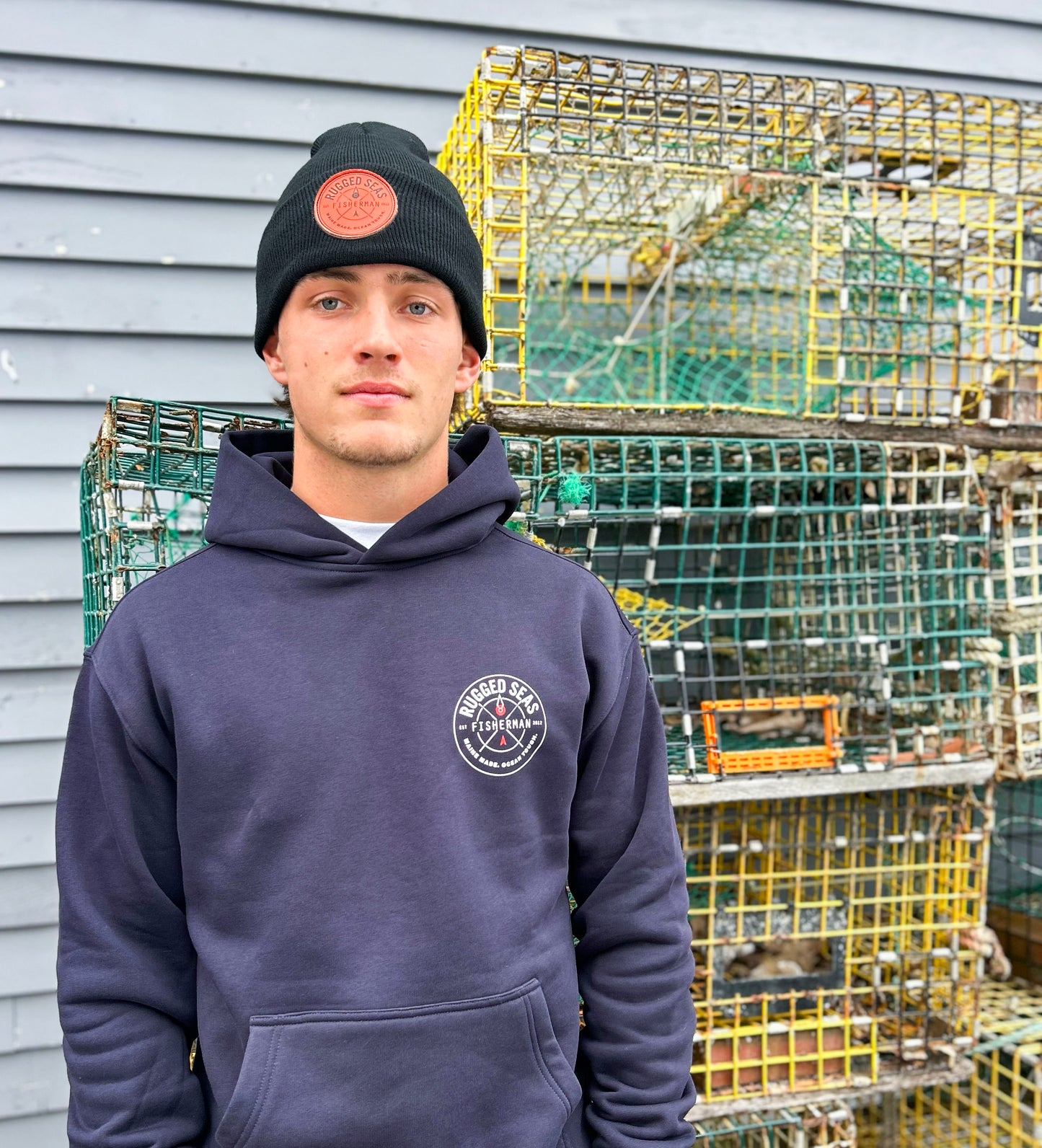 A young man wearing a Rugged Seas Navy Hoodie and a black beanie stands in front of stacked lobster traps beside a gray clapboard wall. by Sea Bags