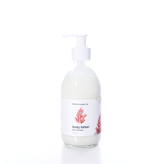 Shore Soap Co. Mermaid Kisses Eco-Friendly Body Lotion in a clear glass pump bottle with a white label featuring a red coral design, photographed on a white background. This natural body lotion provides nourishing care in an elegantly designed, eco-friendly package. by Sea Bags