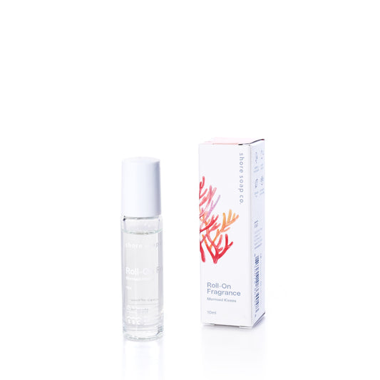 A clear roll-on fragrance bottle labeled Mermaid Kisses Eco-Friendly Roll-On Fragrance by Shore Soap Co. stands beside its white box adorned with coral illustrations on a white background, capturing the fresh and alluring essence characteristic of this beloved scent. by Sea Bags