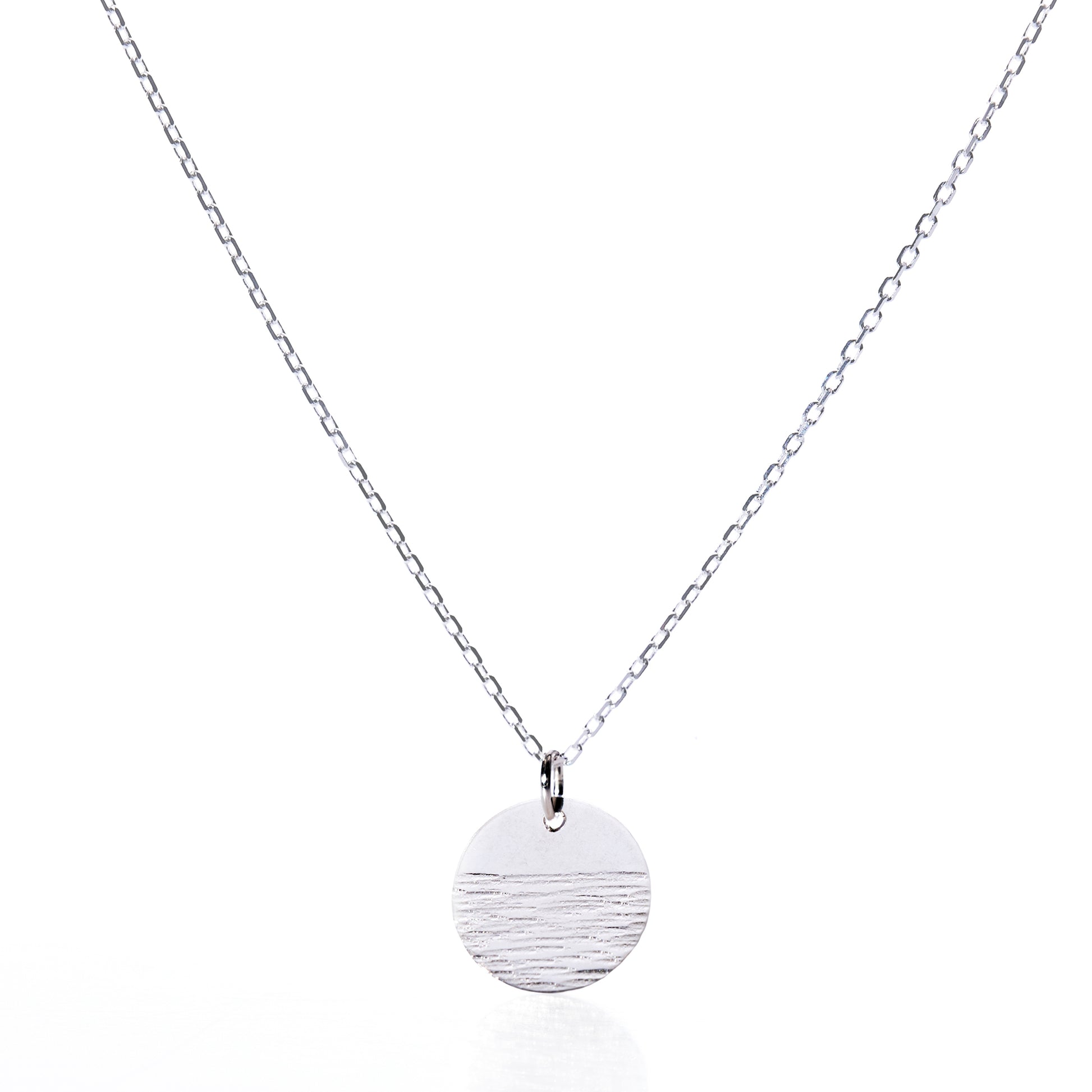 Elevate your jewelry collection with the Pendant Necklace - Horizon Silver by Maine Yoga Jewelry: a refined silver chain showcasing a round, textured silver pendant accented with a distinctive horizontal line detail, beautifully presented against a white background. by Sea Bags