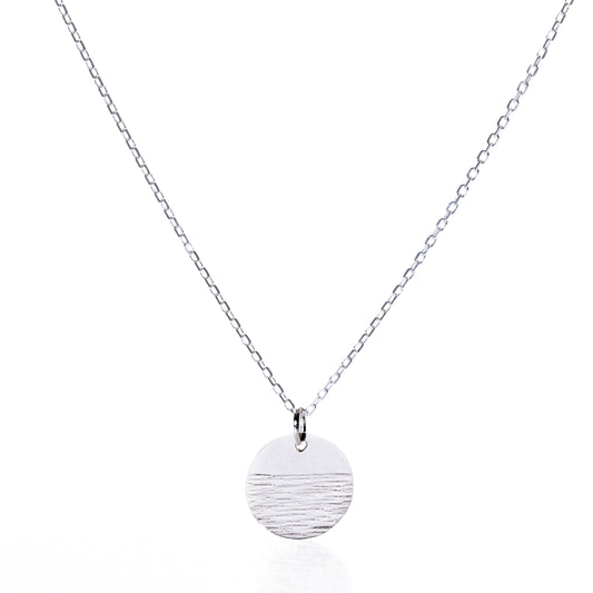 Elevate your jewelry collection with the Pendant Necklace - Horizon Silver by Maine Yoga Jewelry: a refined silver chain showcasing a round, textured silver pendant accented with a distinctive horizontal line detail, beautifully presented against a white background. by Sea Bags