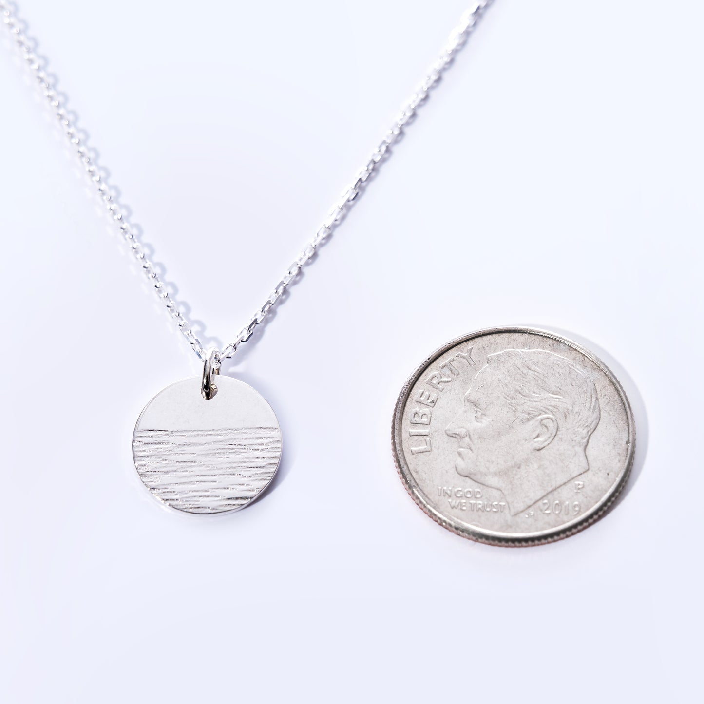 The Pendant Necklace - Horizon Silver by Maine Yoga Jewelry—featuring a round, engraved pendant in a sophisticated silver finish—rests beside a US dime to show its size. by Sea Bags