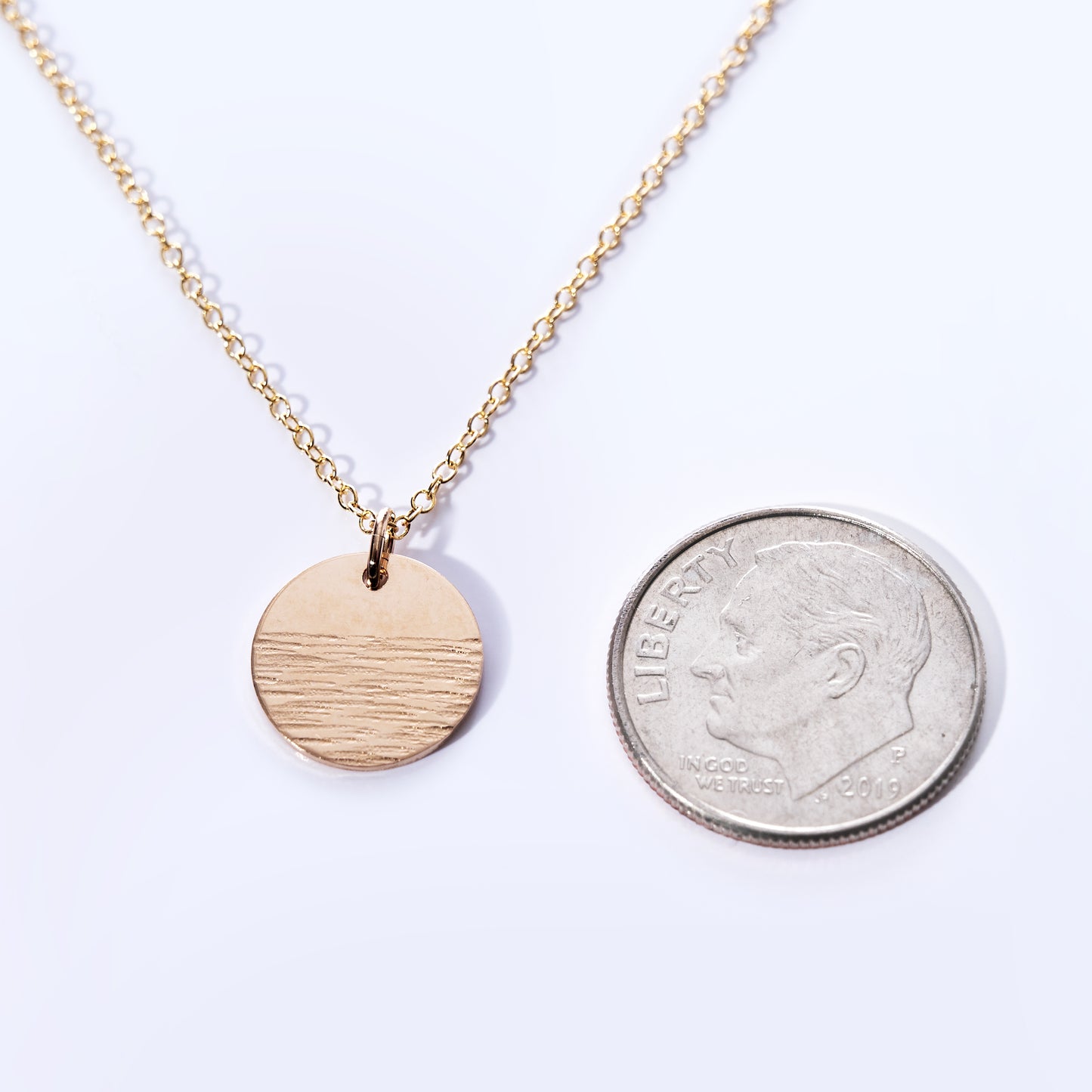 The Pendant Necklace - Horizon Gold by Maine Yoga Jewelry features a round, textured gold pendant on a delicate gold chain, displayed beside a U.S. dime for size comparison on a white background—an elegant piece ideal for enhancing any jewelry collection. by Sea Bags