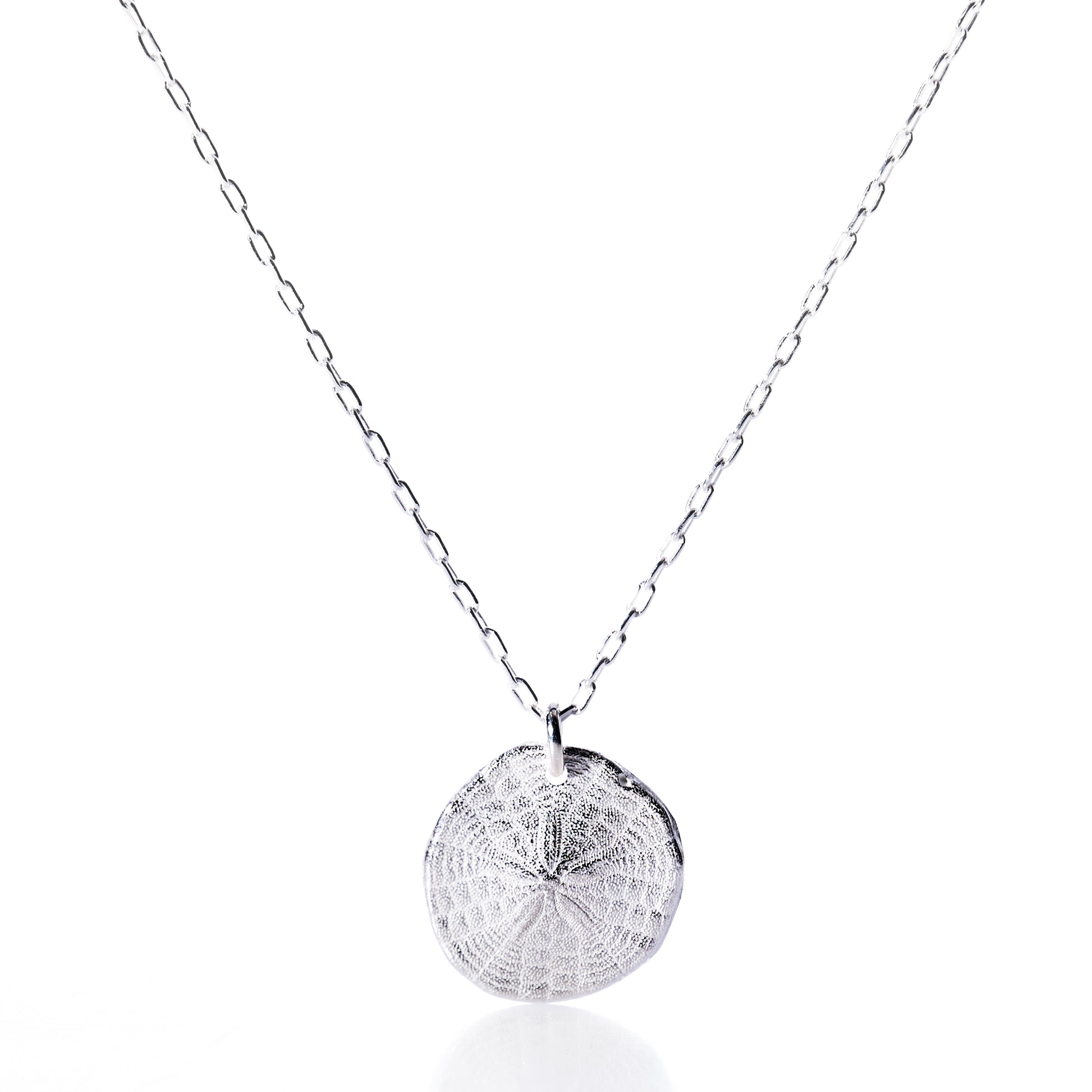 Pendant Necklace - Sand Dollar Silver by Maine Yoga Jewelry features a silver chain necklace with a textured, round sand dollar-inspired pendant, crafted sustainably and evoking the coastal beauty of Maine, shown against a white background. by Sea Bags